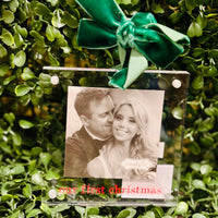 Acrylic ornament with a couple's photo and 'Our First Christmas' text, tied with a green ribbon on a green leafy background.