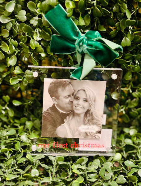 Acrylic ornament with a couple's photo and 'Our First Christmas' text, tied with a green ribbon on a green leafy background.