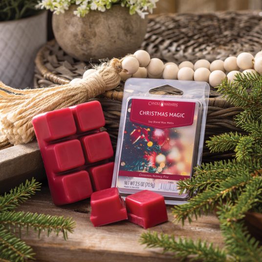 Red wax melts and their packaging labeled 'Christmas Magic' on a wooden surface with greenery and a broom.