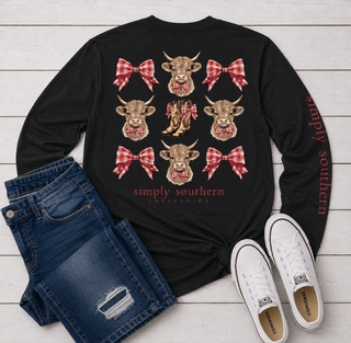 A black long-sleeve graphic tee from Simply Southern Collection. The shirt features a grid of cow illustrations with red and white gingham bows, a red plaid pattern on the sleeve, and 'simply southern' printed on the front and sleeve.