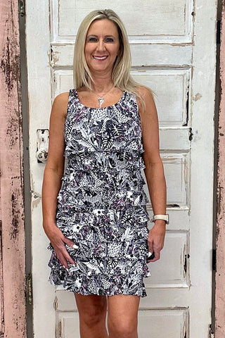 The Lulu-B Cha Cha Dress in the JTBW print features a striking black and white pattern with hot pink accents and hidden animals woven throughout — front and back ruffles, a scoop neckline, and a straight hemline give it a flirty, feminine silhouette that flatters every shape and looks just as stunning at brunch as it does on vacation.