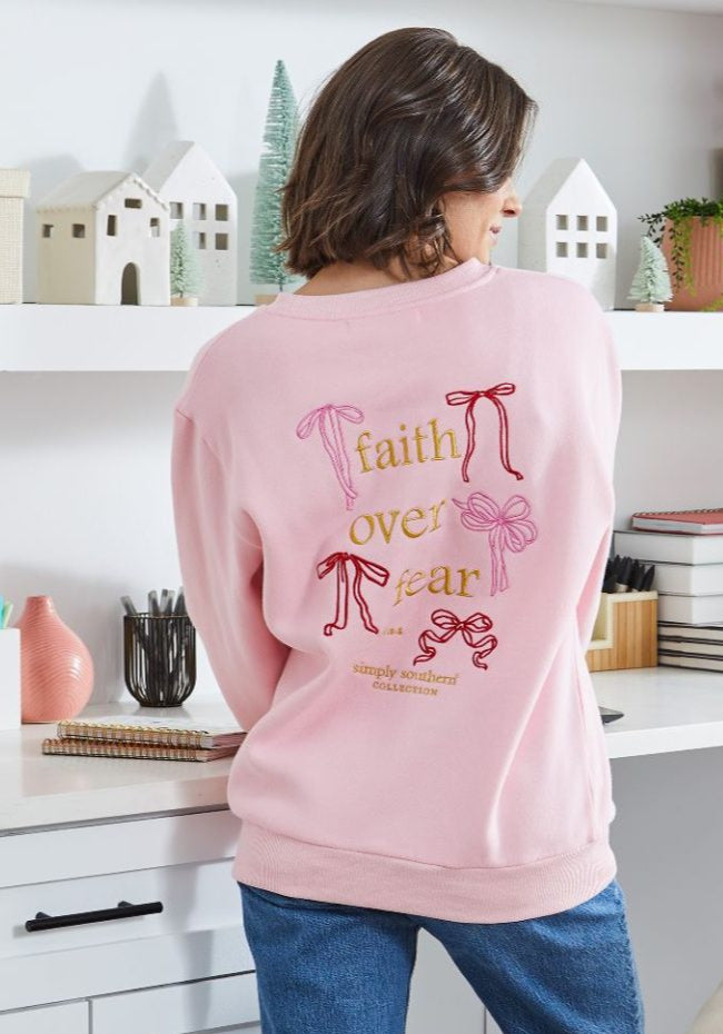 Person wearing a pink sweatshirt with 'faith over fear' text in a home setting