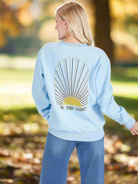 Light blue sweatshirt with a sun design and 'Be the Light' text on the back, worn by a person.