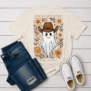 A cream-colored graphic t-shir. The t-shirt features a cartoon ghost wearing a brown cowboy hat, surrounded by orange flowers and green leaves, with the text 'APPARENTLY I HAVE AN ATTITUDE simply southern'.