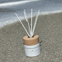 White ceramic jar featuring a wooden cap and bamboo reeds with True Ocean's signature Coastal scent. A beach day fragrance with notes of bergamot, palm leaf, and mandarin. Bringing a carefree vibe of sand, surf, and ocean waves. Packaged in a premium gift box with embossed silver lettering. The perfect beach gift for any occasion.