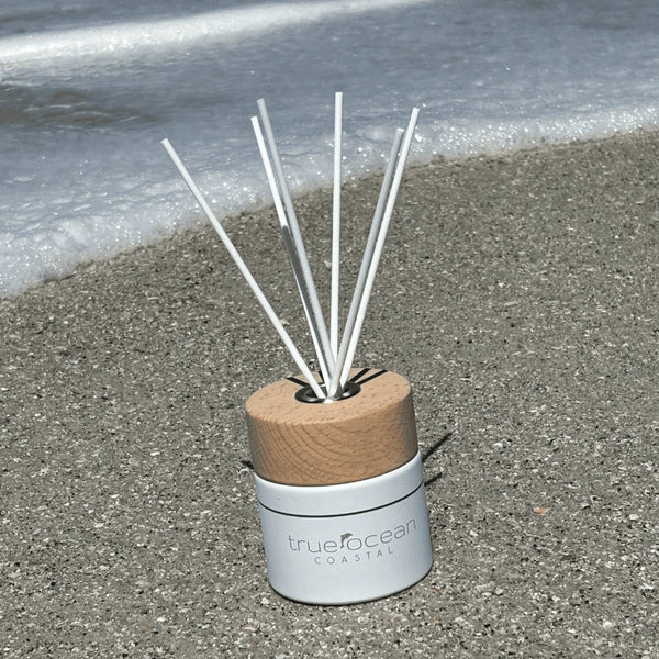 White ceramic jar featuring a wooden cap and bamboo reeds with True Ocean's signature Coastal scent. A beach day fragrance with notes of bergamot, palm leaf, and mandarin. Bringing a carefree vibe of sand, surf, and ocean waves. Packaged in a premium gift box with embossed silver lettering. The perfect beach gift for any occasion.