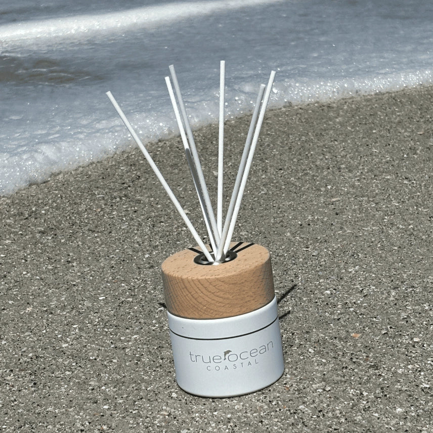 White ceramic jar featuring a wooden cap and bamboo reeds with True Ocean's signature Coastal scent. A beach day fragrance with notes of bergamot, palm leaf, and mandarin. Bringing a carefree vibe of sand, surf, and ocean waves. Packaged in a premium gift box with embossed silver lettering. The perfect beach gift for any occasion.